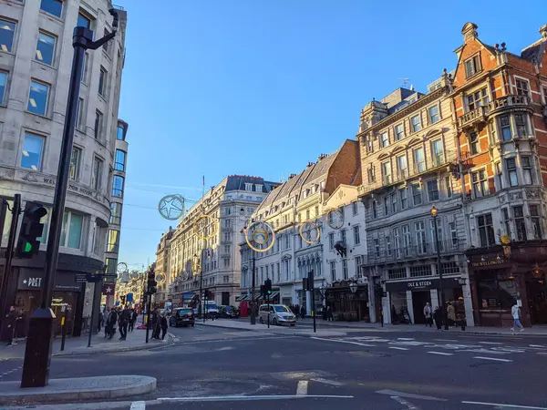 Londra, İngiltere - 05 Kasım 2021: Yayalar yürür ve arabalar, açık mavi gökyüzü altında sıcak öğleden sonra güneşiyle aydınlanan Strand boyunca klasik Londra binalarının ve dükkanlarının önünden geçer