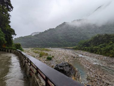 Taroko, Tayvan - 26 Kasım 2022: Bulutlu bir günde sisle kaplanmış yemyeşil dağların altındaki vadide parmaklıkları olan boş bir yürüyüş yolunun yanında kayalık nehir yatağı ve akan su