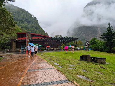 Taroko, Tayvan - 26 Kasım 2022: Şemsiyeli ziyaretçiler, yemyeşil çimenler ve sisli dağ tepeleriyle çevrili modern Buluowa Servis İstasyonu 'na doğru ıslak tuğla bir patika boyunca yürüyorlar