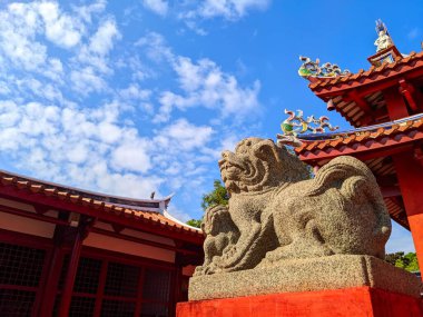 Tainan, Tayvan - 02 Aralık 2022: Taş aslan heykeli Tainan Konfüçyüs Tapınağı 'nın önünde kırmızı sütunları ve parlak mavi gökyüzünün altında süslü çatı detaylarıyla nöbet tutuyor.