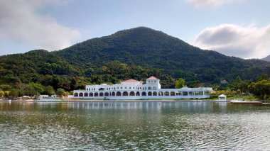 Hong Kong - 28 Şubat 2021: Kemerli ve kırmızı çatılı Göl Evi adında beyaz bir bina, Egret Gölü 'nün yanında bulutlu bir gökyüzünün altında yemyeşil bir dağı yansıtıyor.