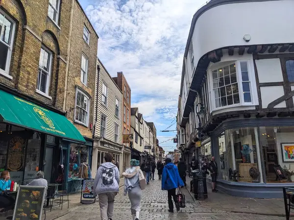 Canterbury, İngiltere - 19 Nisan 2025: Yayalar, bulutlu bir gökyüzü altında dükkanlar ve geleneksel mimari ile sıralanmış tarihi kaldırım geçidi boyunca yürürler ve şehrin canlı atmosferini yakalarlar