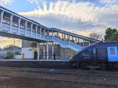 Canterbury, İngiltere - 19 Nisan 2025: Bir Güneydoğu Javelin treni Canterbury Batı İstasyonu platformuna ulaşırken, insanlar dramatik bulutlu bir gökyüzü altında yaya üstgeçidinde yürüyor.