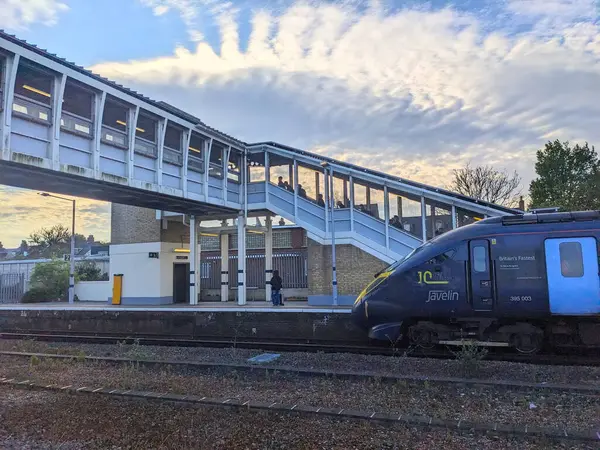 Canterbury, İngiltere - 19 Nisan 2025: Bir Güneydoğu Javelin treni Canterbury Batı İstasyonu platformuna ulaşırken, insanlar dramatik bulutlu bir gökyüzü altında yaya üstgeçidinde yürüyor.