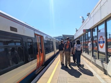 Londra, İngiltere - Nisan 06 2025: Londra Metro istasyonunda açık mavi gökyüzü altında bekleyen güneşli bir platform boyunca yürüyen gezginler ve banliyö yolcuları