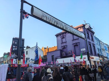 Londra, İngiltere - 11 Ocak 2025: Hareketli bir kalabalık Camden Town 'da açık mavi bir gökyüzünün altındaki Inverness Caddesi Market levhasının altında canlı mağaza cephelerinin ve market tezgahlarının önünden geçiyor