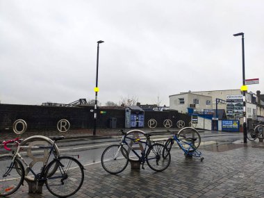 Londra, İngiltere - 01 Aralık 2024: Bisikletler, Manor Park Elizabeth Line İstasyonu 'nun dışındaki bir yaya geçidinin yanındaki ıslak bir kaldırımda, gri bulutlu gökyüzünün altında park halindedir.