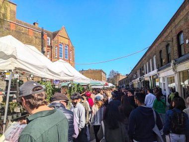 Londra, İngiltere - Nisan 06 2025: Büyük bir müşteri ve ziyaretçi kalabalığı Kolombiya Yolu Çiçek Pazarı 'nda hareketli bir şekilde yürüyor ve santral ve tarihi tuğla binalar satan tedarikçi çadırlarıyla sıralanmış durumda