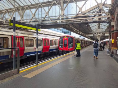 Londra, İngiltere - 26 Nisan 2025: Yolcular ve bir demiryolu işçisi platformda dururken, kırmızı bir Londra metro treni Hammersmith İstasyonu 'na yanaşıyor.