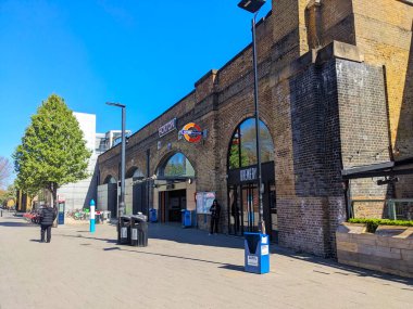 Londra, İngiltere - Nisan 06 2025: Açık mavi gökyüzünün altında tuğla kemerli Hoxton Overground istasyon binasının girişi, kaldırımda yürüyen yayaların ve park halindeki bisikletlerin yer aldığı