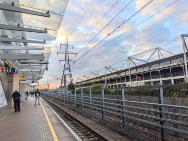 Londra, İngiltere - 25 Nisan 2024: Gümrük Evi istasyonu Elizabeth Line platformunda güneş batarken, bulutlu bir gökyüzü altında bir elektrik direği ve Londra Excel Center 'da bekleyenler