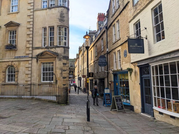 Bath, UK - January 30 2025: Pedestrians stroll along a cobblestone street lined with classic Georgian architecture and traditional shops like Sally Lunns in the historic city under a bright sky