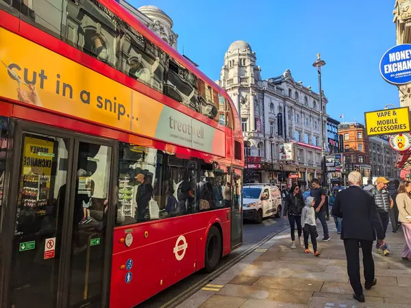 Londra, İngiltere - 10 Nisan 2025: Meşgul Shaftesbury Bulvarı boyunca yayalar ve mavi gökyüzü altında tarihi binalarla dolu ünlü kırmızı çift katlı bir otobüs sürüyor