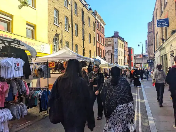 Londra, İngiltere - Nisan 06 2025: Müşteriler ve satıcılar Brick Lane 'deki canlı sokak pazarını açık mavi gökyüzü altında giyim ve mal satan tezgahlarla doldurdular
