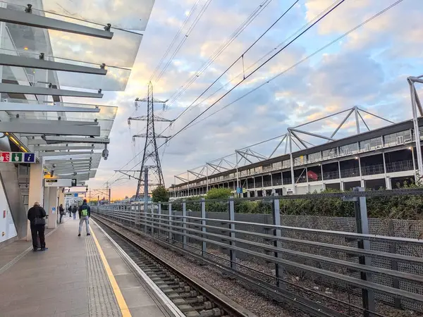 Londra, İngiltere - 25 Nisan 2024: Gümrük Evi istasyonu Elizabeth Line platformunda güneş batarken, bulutlu bir gökyüzü altında bir elektrik direği ve Londra Excel Center 'da bekleyenler