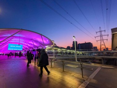 Londra, İngiltere - 13 Şubat 2023: Banliyö sakinleri, Royal Rıhtımı 'ndaki Gümrük Evi İstasyonu' nda şehir binaları ve bir pilon ile canlı bir alacakaranlık gökyüzüne karşı modern aydınlatılmış yaya geçidi boyunca yürüyor