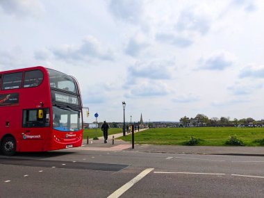 Londra, İngiltere - 30 Nisan 2023: Kırmızı bir posta arabası, bulutlu bir günde Blackheath 'de büyük bir yeşil parkın önünden geçerek yürüyen ve scooter kullanan insanlarla cadde boyunca ilerliyor
