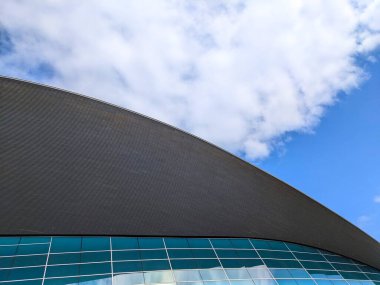 Londra, İngiltere - 17 Nisan 2023: Düşük açılı soyut görüş Londra Sucul Merkezi 'nin modern eğimli çatı ve mavi cam cephesini bulutlu bir gökyüzü altında görüntülemek