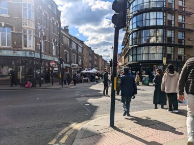 Londra, İngiltere - Nisan 03 2022: Yayalar, Brick Lane 'de günışığıyla aydınlanan bir sokak köşesinden geçerek, geleneksel tuğla binaların parçalı bulutlu bir gökyüzü altında modern cam mimari ile tezat oluşturuyorlar.
