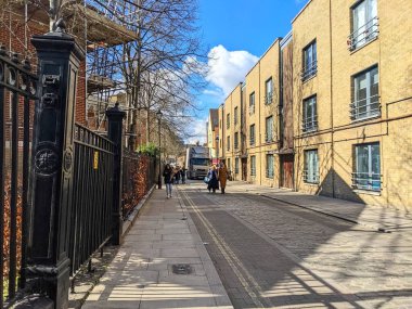 Londra, İngiltere - 3 Nisan 2022: Yayalar, günışığıyla aydınlanan kaldırımlar ve modern tuğla konut binaları ve mavi gökyüzünün altında süslü bir demir çit ile kaplı kaldırımlar boyunca yürüyorlar