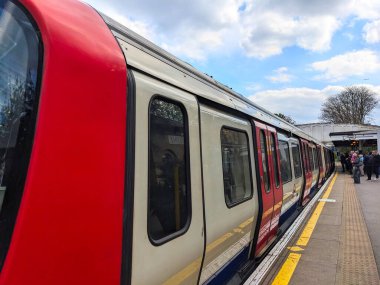 Londra, İngiltere - 26 Nisan 2024: Klasik bir kırmızı, beyaz ve mavi Londra Metro treni, parçalı bulutlu bir gökyüzü altında uçmak için bekleyen Kew Garden İstasyonu açık hava platformuna ulaşır.