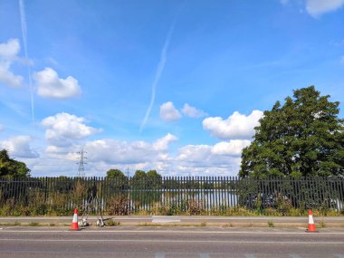 Londra, İngiltere - 21 Eylül 2021: Walthamstow Bataklıkları, parlak mavi gökyüzü altında canlı yeşil ağaçlar ve uzak elektrik direkleriyle çevrili sakin bir yol manzarası sergileniyor