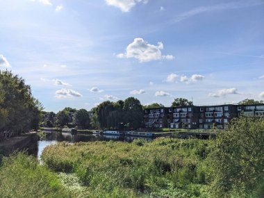 Londra, İngiltere - 21 Eylül 2021: Narrowboat 'lar sakin Lea Nehri' nde konut binaları ve açık mavi gökyüzü altında güneşli yemyeşil kamışlarla birlikte süzülür