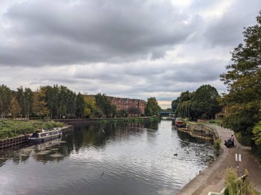 Londra, İngiltere - 13 Ekim 2021: Lea Nehri 'nin sessiz bir uzantısı, su boyunca kenetlenmiş yüzen teknelere, geleneksel tuğla binalara ve bulutlu bir sonbahar gökyüzünün altında yemyeşil nehir kenarındaki ağaçlara sahiptir.