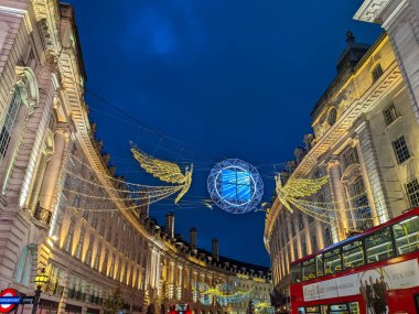 Londra, İngiltere - 21 Aralık 2021: Noel Meleğinin Şenlikli Ruhu Regent Caddesi 'nin büyük mimarisinin üzerinde alacakaranlıkta kırmızı çift katlı bir otobüs geçerken ışıldıyor