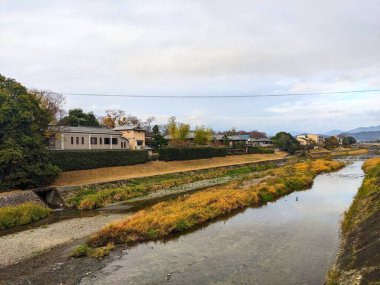 Kyoto, Japonya - 15 Aralık 2023: Kamo Nehri 'nin sakin ve bulutlu bir gökyüzü altında uzak dağlarla meskun Japon evleri ve kış yapraklarının yanından geçerken sakin bir görüntüsü