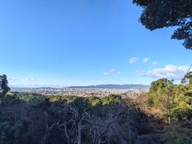 Fushimi, Japonya - 17 Aralık 2023: Kyoto 'nun uzak dağlara uzanan kentsel manzarasının geniş panoramik görünümü, açık bir gökyüzü altında Inari Dağı' nın yüksek ormanlı bir perspektifinden görülüyor