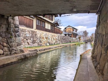 Shiga, Japonya - 20 Aralık 2023: Bulutlu bir günde tarihi Hachiman-bori kanalı boyunca uzanan ve taştan duvarlar ve geleneksel ticaret evleriyle çevrili bir köprü altından manzara