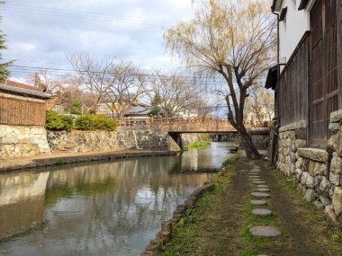 Shiga, Japonya - 20 Aralık 2023: Geleneksel taş duvarları, ahşap bir köprüsü ve suların yüzeyindeki çıplak kış ağaçlarının yansımaları ile tarihi Hachiman-bori kanalının sakin bir görüntüsü