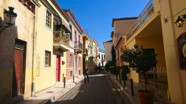 Atina, Yunanistan - 29,3,2018: Salgından önceki yaz güneşli açık mavi bir gökyüzünün altında, sakin bir sokakta renkli konutlarla yürüyen yayalar