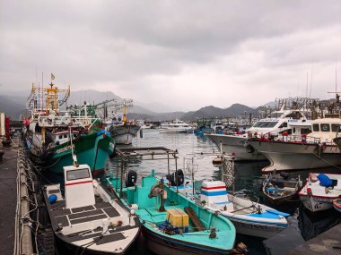 New Taipei, Tayvan - 24.12.2023: Balıkçı tekneleri ve yatlar Shenao Limanı 'nın kenarında, bulutlu bir gökyüzünün altında, arkada sislerin arasında gizlenmiş bir dağ silsilesiyle demirli.