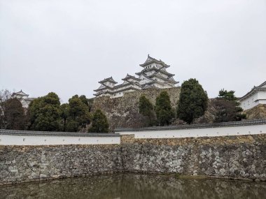 Himeji, Japonya - 12.19.2023: Geniş Üç Ülke Hendeği 'nin arkasındaki Himeji Şatosu' nun ana kalesi ve önü bulutlu bir gökyüzünün altında iyi korunan ağaçlarla çevrili duvarlar.