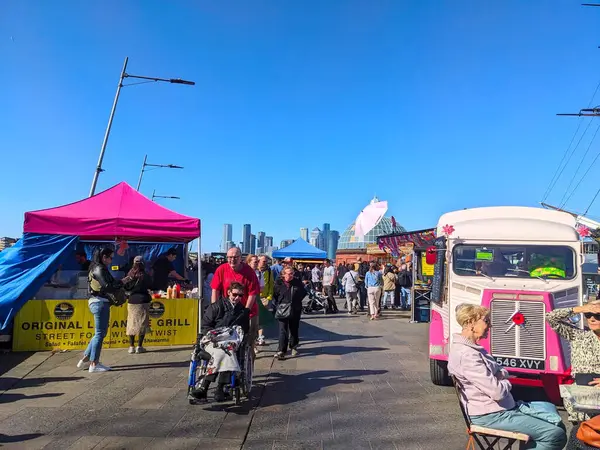 London, UK - April 05 2025: The lively outdoor Cutty Sark Street Food Market is bustling with diverse people browsing food stalls and enjoying a sunny day with the city skyline in the background