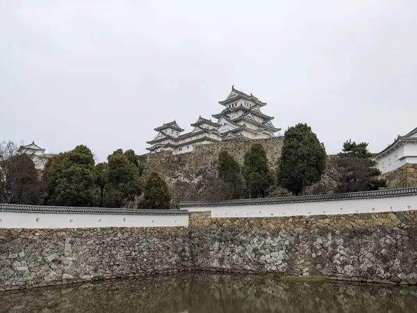 Himeji, Japonya - 12.19.2023: Geniş Üç Ülke Hendeği 'nin arkasındaki Himeji Şatosu' nun ana kalesi ve önü bulutlu bir gökyüzünün altında iyi korunan ağaçlarla çevrili duvarlar.