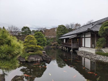 Himeji, Japonya - 12.19.2023: Turistler olmadan bulutlu bir gökyüzünün altında Koko-en 'de ağaçlarla çevrili barışçıl bir gölet üzerinde ahşap bir gözlem güvertesi bulunan geleneksel bir ev
