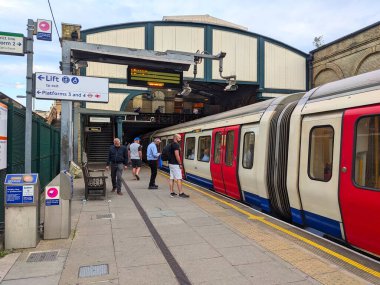 Londra, İngiltere - 07.31.2024: Batı Brompton İstasyonu 'ndaki platforma binmek için bekleyen yolcular ve arkada bulutlu saatler sırasında bulutlu bir gökyüzünün altında iki pembe kart okuyucusu