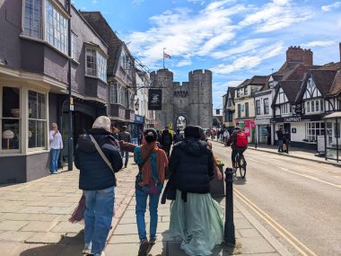 Canterbury, UK - April 19 2025: Pedestrians stroll along the pavement of St. Dunstans Street and a cyclist rides past historic Westgate Towers and traditional shops under a bright, partly cloudy sky