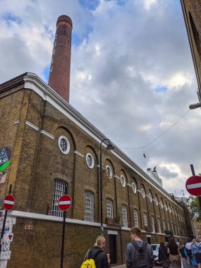 Londra, İngiltere - 09.26.2021: Bulutlu mavi gökyüzünün altında tarihi bir bacayla Kazan Evi 'nin dışındaki işlek tuğla arazisinde yürüyen yayalar