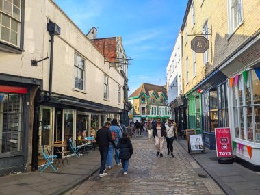 Canterbury, UK - April 19 2025: People walk past historic buildings housing independent shops and businesses on the traditional cobbled street Burgate in the city centre under a sunny blue sky