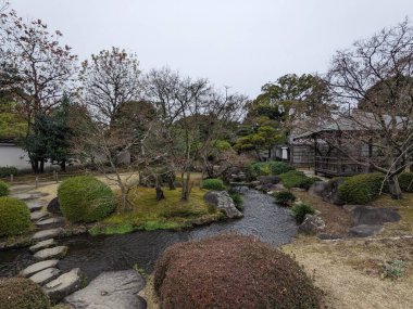 Himeji, Japonya - 12.19.2023: Bulutlu bir gökyüzünün altında manikürlü ağaçlarla çevrili Koko-en 'de geleneksel bir Japon bahçesindeki ahşap bir çadırın yanındaki derenin üzerindeki taş patika