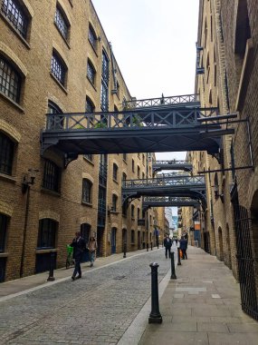 Londra, İngiltere - 05.13.2024: Shad Thames caddesinde yürüyen ve konuşan insanlar dönüştürülmüş depolar ve fayanslı zemin ve zemin kattaki mağazalar üzerindeki yükseltilmiş yürüyüş yolları bulutlu bir gökyüzü altında