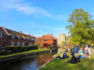 Canterbury, İngiltere - 19 Nisan 2025: Westgate Gardens 'ta güneşli bir bahar öğleden sonrasında tarihi binalar ve Westgate Kuleleri' nin arka planında nehir kenarındaki çimlerde dinlenen insanlar