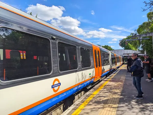 Londra, İngiltere - 06.19.2024: Bir Londra Overground treni Kew Gardens İstasyonu 'ndaki platforma yaklaşırken yolcular açık mavi gökyüzü altında bulutlar ve güneş ışığıyla birlikte yaz aylarında trene binmeye hazırdır.