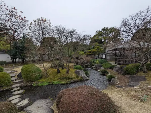 Himeji, Japonya - 12.19.2023: Bulutlu bir gökyüzünün altında manikürlü ağaçlarla çevrili Koko-en 'de geleneksel bir Japon bahçesindeki ahşap bir çadırın yanındaki derenin üzerindeki taş patika