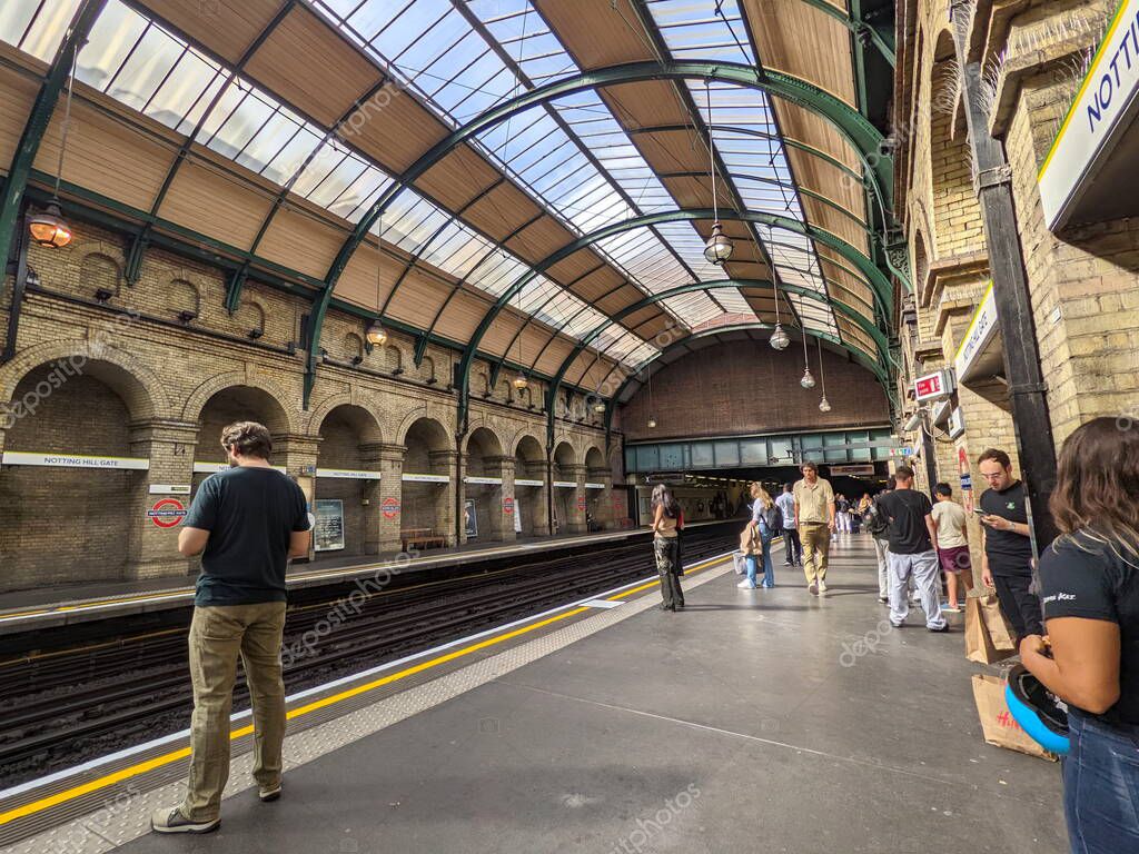 London, UK - 2022.09.04: Passengers waiting for a train on the District and Circle Line platform at Notting Hill Gate on a cloudy day under a curved glass ceiling with hanging lamps and brick arches