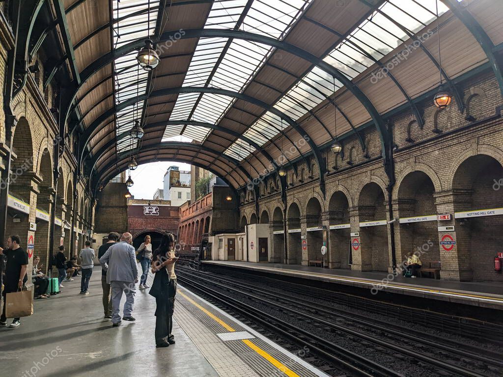 London, UK - 2022.09.04: Passengers waiting for a train on the District and Circle Line platform at Notting Hill Gate on a cloudy day under a curved glass ceiling with hanging lamps and brick arches